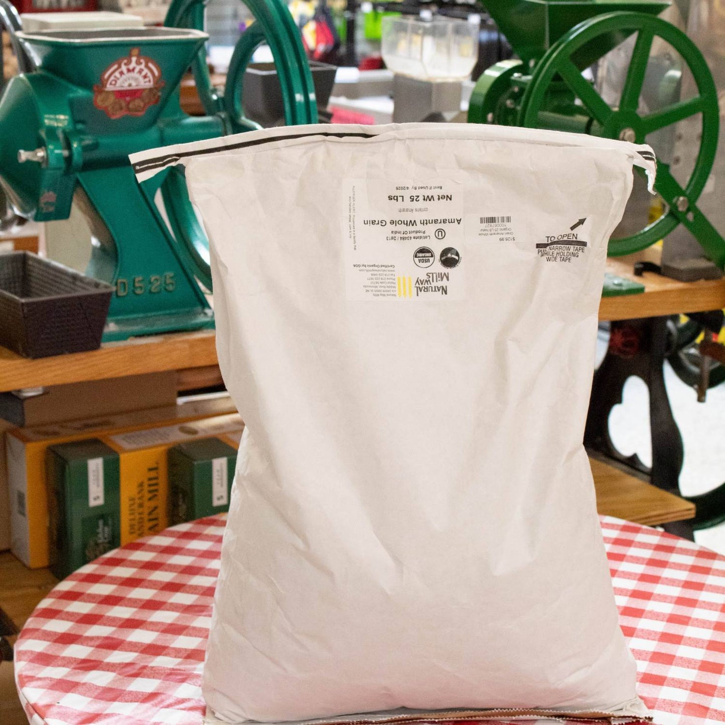 Tall white paper sack of whole amaranth grain weighing 25 pounds, set on a red and white checkered table with vintage grain mills and kitchen tools in the background, ideal for pantry and food collections.