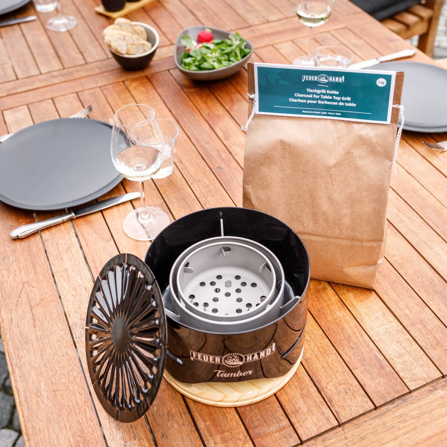Black Tamber tabletop grill with an open vented lid next to a brown paper bag of charcoal, set on a wooden outdoor table arranged with plates, glassware, and fresh salad, featuring grilling essentials for Stoves, Grilling, Best Sellers, and Promo Eligible collections.