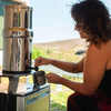 Woman filling a cup with filtered water from a stainless steel gravity water filter outdoors, featuring a durable design ideal for camping, emergency supplies, and farm use.