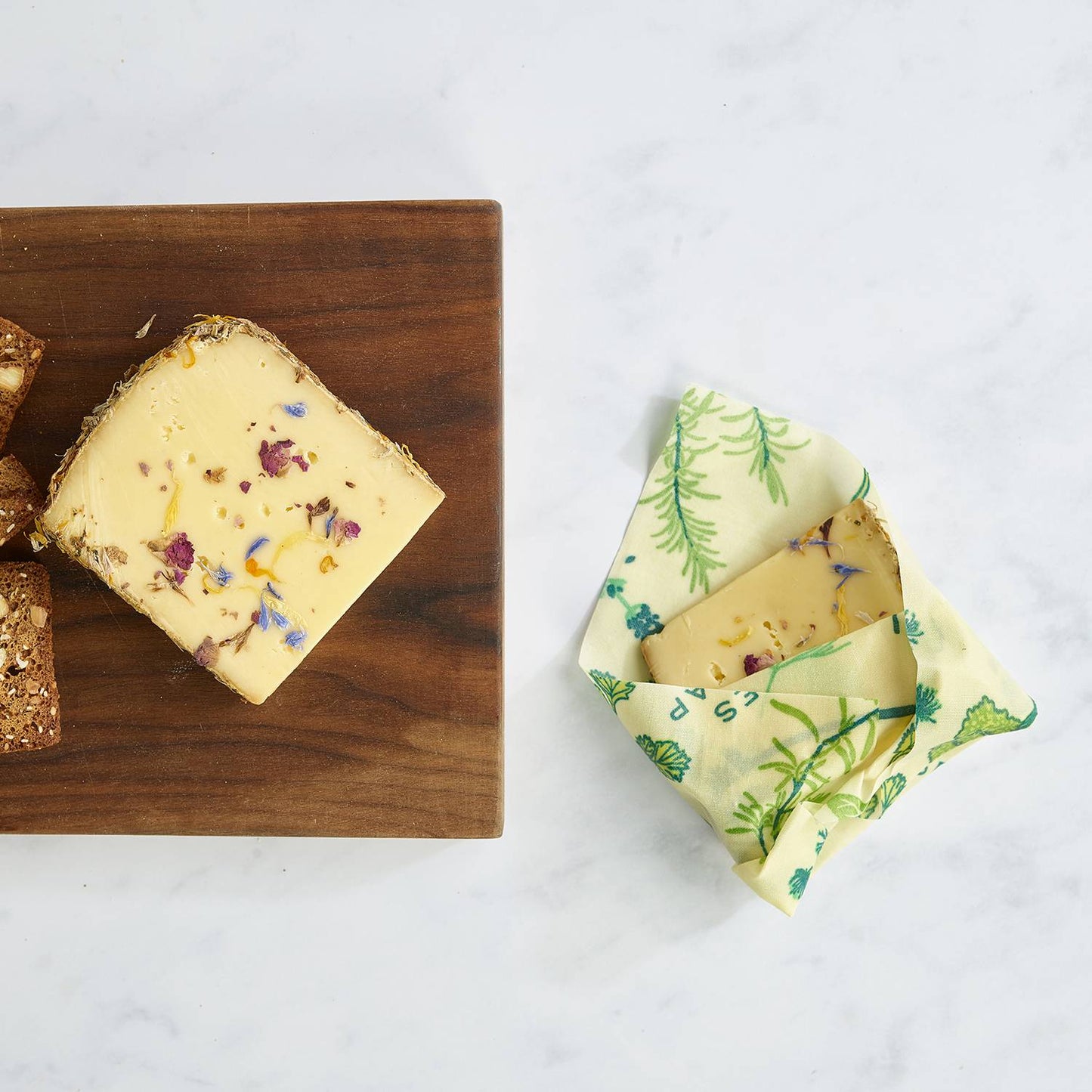 Small square of handmade beeswax wrap with green botanical print securing a piece of artisanal cheese on white marble, part of Kitchen & Food and USA Made collections.