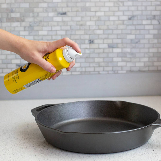 Hand spraying yellow canola oil seasoning spray onto a seasoned cast iron skillet, ideal for Kitchen & Food, Cookware, Cast Iron, Best Sellers, and Promo Eligible collections.