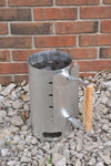 Aluminized steel charcoal starter with wooden handle standing on gravel against a brick wall, designed for quick briquet ignition, ideal for camping and farm tool collections.