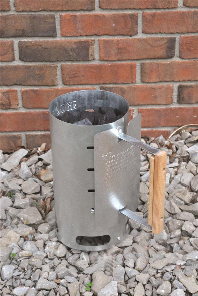 Aluminized steel charcoal starter with wooden handle standing on gravel against a brick wall, designed for quick briquet ignition, ideal for camping and farm tool collections.