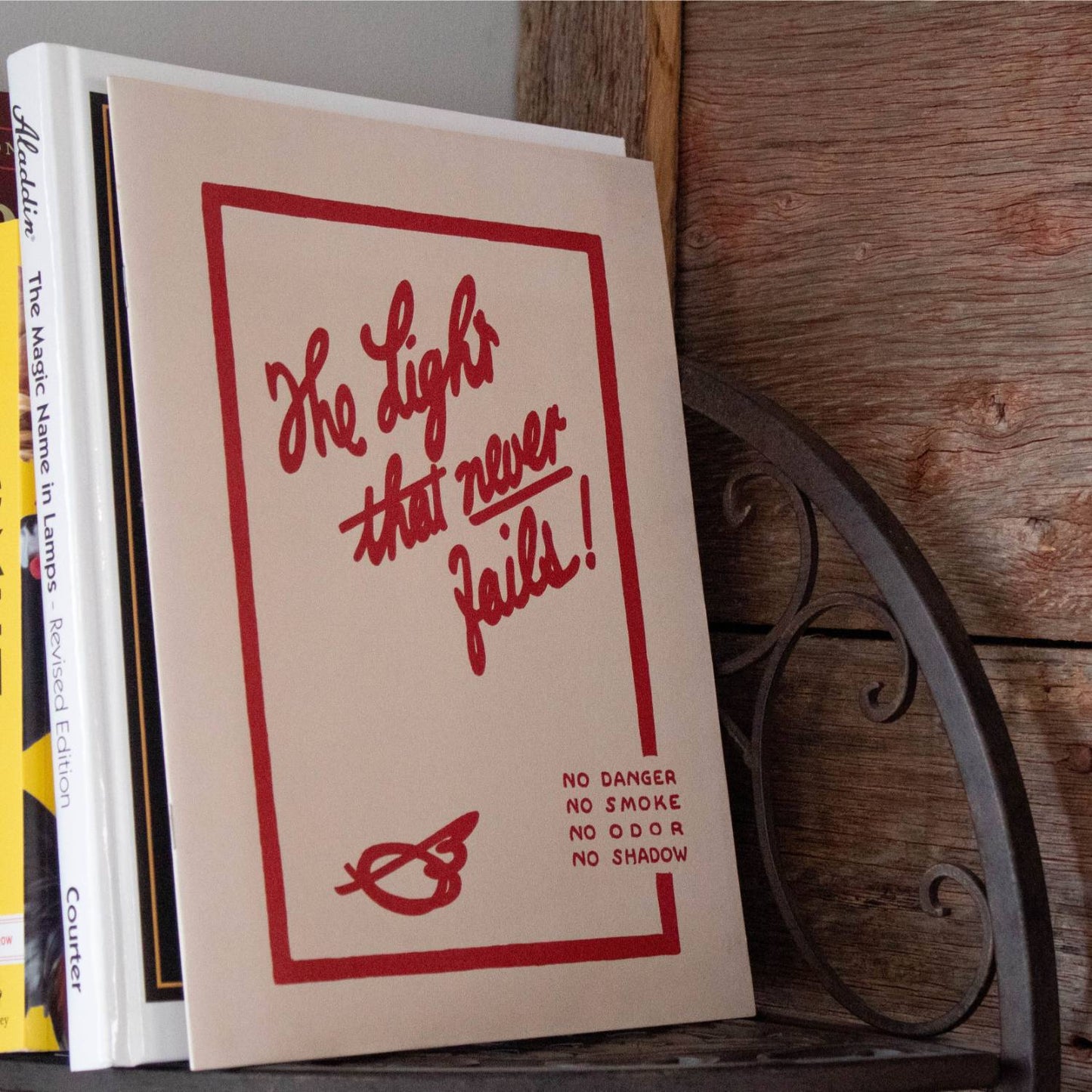 Paperback booklet with red handwritten-style text reading "The Light that never fails!" alongside safety assurances, resting on a rustic wooden shelf with curved iron supports, suitable for Heritage and Lifestyle Books and Toys, Books & Games collections.