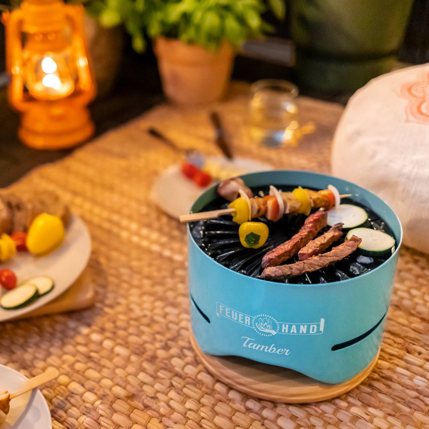 Small turquoise tabletop charcoal grill with curved vented sides cooking skewered vegetables and sliced meat, placed on a wooden trivet and woven mat, surrounded by cozy outdoor dining elements in the Farm, Garden & Tool and Grilling collections.