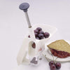 White clamp-on cherry pitter with a spring-loaded plunger, shown with fresh cherries loaded in the funnel and pitted cherries collected below, alongside a slice of cherry pie on a plate, suitable for Kitchen & Food, Specialty Equipment, Peelers and Pitters, and Best Sellers collections.