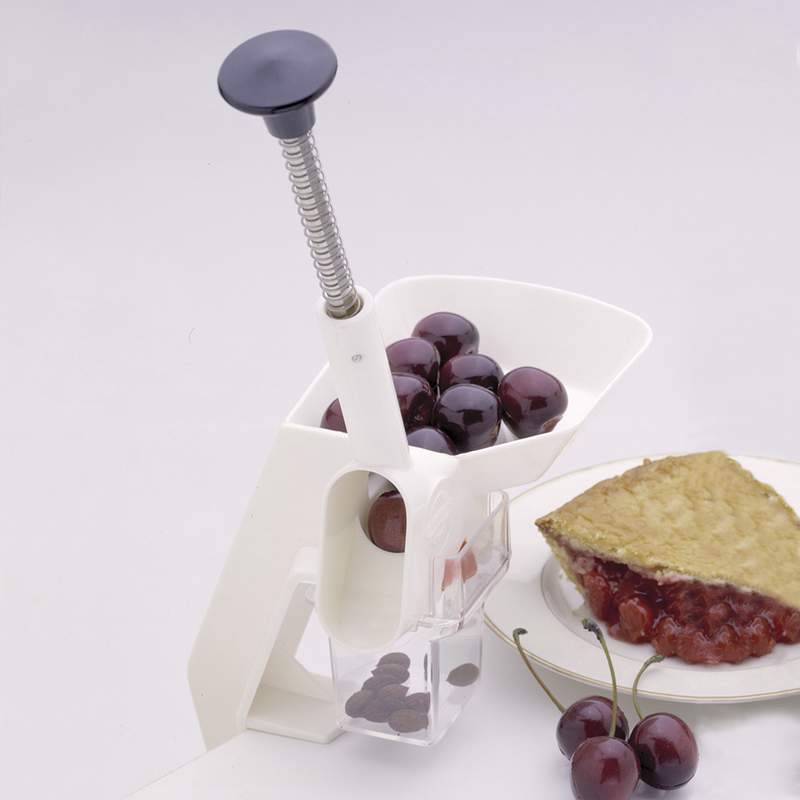 White clamp-on cherry pitter with a spring-loaded plunger, shown with fresh cherries loaded in the funnel and pitted cherries collected below, alongside a slice of cherry pie on a plate, suitable for Kitchen & Food, Specialty Equipment, Peelers and Pitters, and Best Sellers collections.
