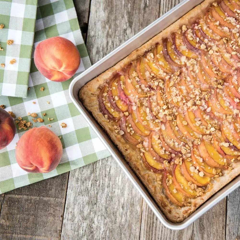 High-sided aluminum sheet pan filled with a freshly baked peach dessert topped with nuts, set on a rustic wooden table with fresh peaches and a green checkered cloth, ideal for kitchen and baking supplies collections.