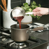 Hand pouring liquid from a heavy-duty stainless steel measuring pan with internal markings into a saucepan on a gas stove, ideal for Kitchen & Food, Cookware, and Baking Supplies collections.