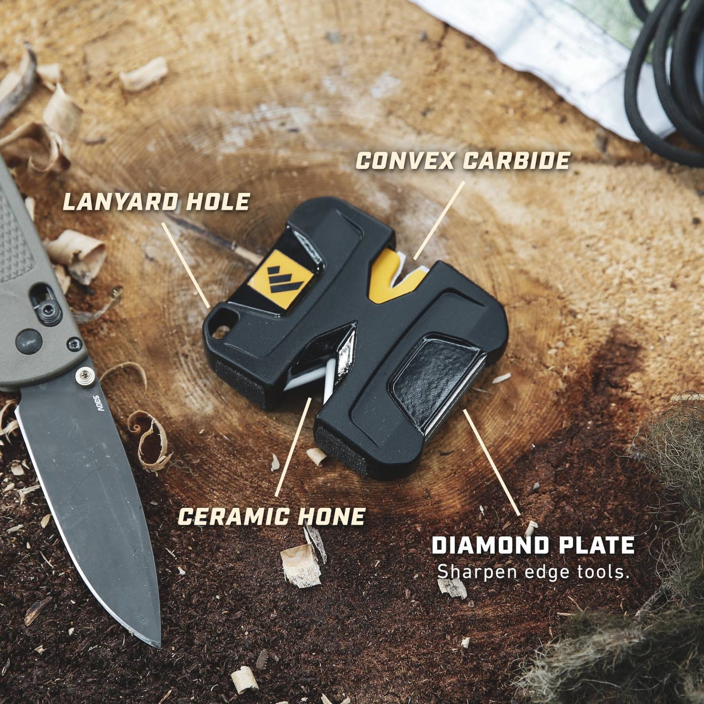 Compact black pocket-sized knife sharpener showing labeled features including convex carbide, ceramic hone, diamond plate, and lanyard hole, displayed on a wooden surface with a folding knife nearby.