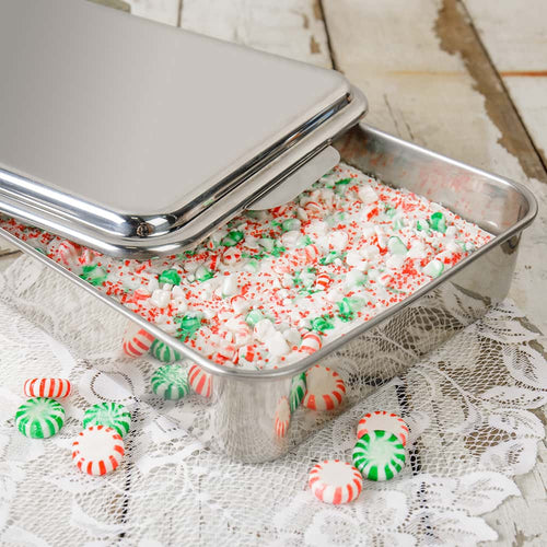 Rectangular stainless steel cake pan with a raised lid partially open, holding a festive dessert topped with green and red peppermint candies and small marshmallows, placed on a lace tablecloth.