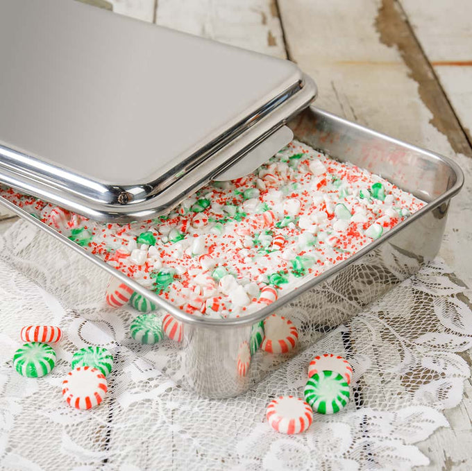 Rectangular stainless steel cake pan with a raised lid partially open, holding a festive dessert topped with green and red peppermint candies and small marshmallows, placed on a lace tablecloth.