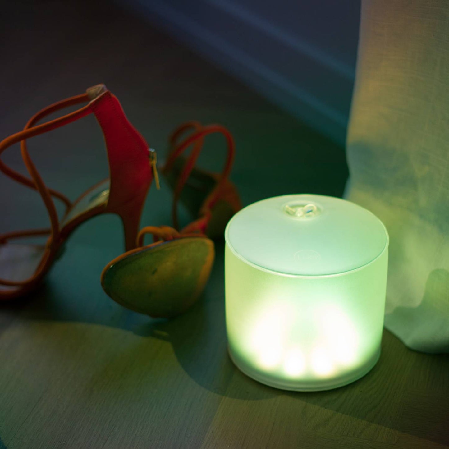 Soft green glowing Luci Color Essence solar light casting ambient illumination on a wooden floor next to a pair of red high-heeled sandals, ideal for eco-friendly Lamps & Lighting and cozy indoor settings.