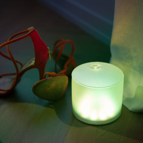 Soft green glowing Luci Color Essence solar light casting ambient illumination on a wooden floor next to a pair of red high-heeled sandals, ideal for eco-friendly Lamps & Lighting and cozy indoor settings.