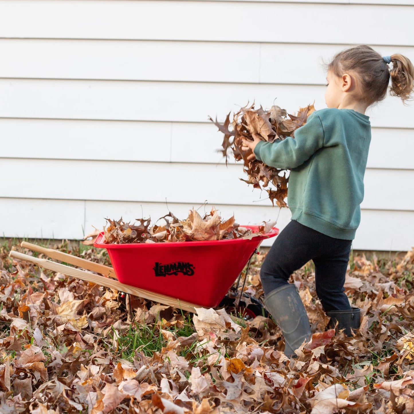 Child playing outdoors with a red handcrafted kids wheelbarrow by Lehman’s, helping collect autumn leaves, in a yard setting promoting outdoor play and garden fun collections.