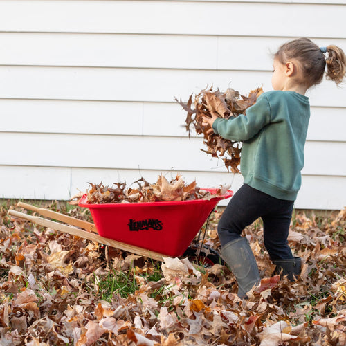 Child playing outdoors with a red handcrafted kids wheelbarrow by Lehman’s, helping collect autumn leaves, in a yard setting promoting outdoor play and garden fun collections.