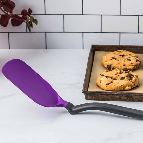 Purple flexible spatula with a black handle resting on a white countertop near a baking sheet with two chocolate chip cookies, ideal for Kitchen & Food, Kitchen Gadgets, and Best Sellers collections.