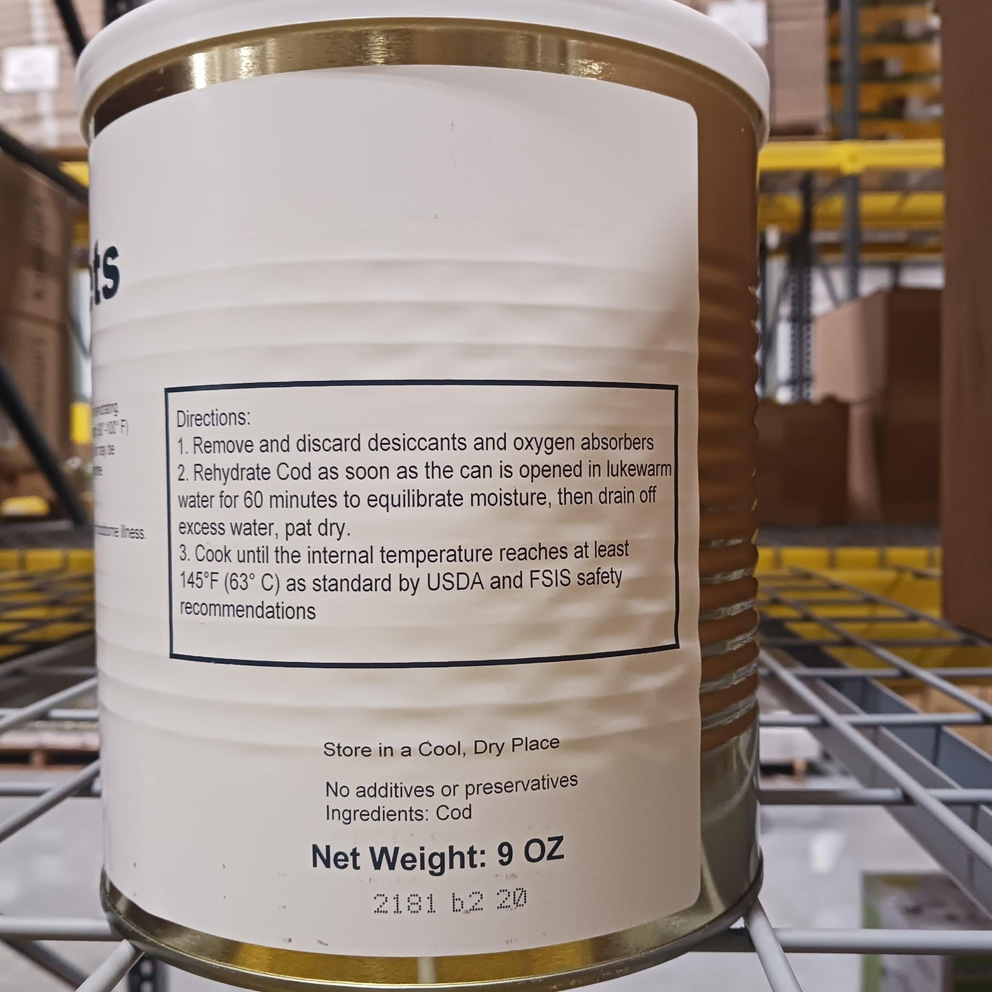 Close-up of a canned freeze-dried cod product label showing preparation instructions, storage advice, ingredient listing, and net weight in a warehouse setting behind shelving.
