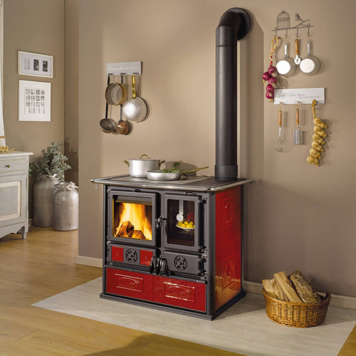 Red and black ceramic wood cook stove with fire burning inside, featuring panoramic enameled oven, vitrified cast-iron top, cooking pots, and a wood basket, staged in a cozy kitchen setting.