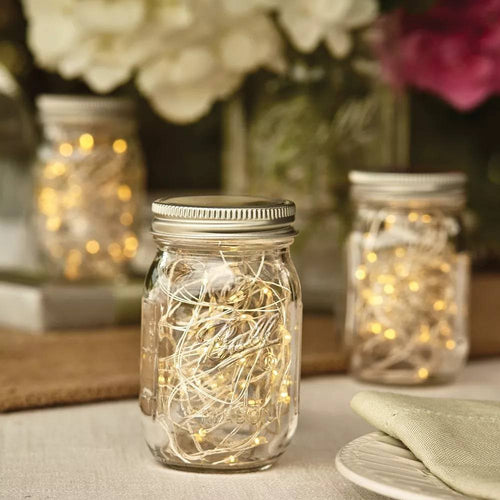 Clear mini glass jar with metal lid filled with warm string lights, arranged on a table with blurred jars and floral decor in the background, fitting Home & Decor and USA Made collections.