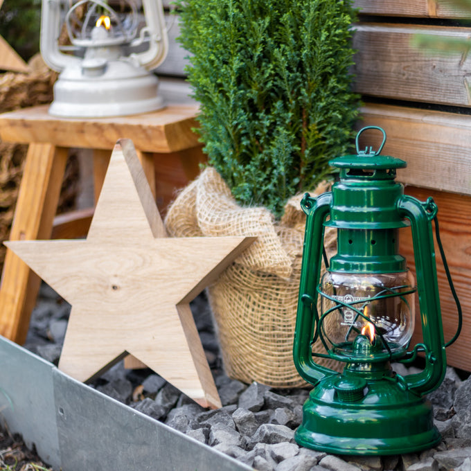 Handcrafted green galvanized oil lantern with heat-resistant SUPRAX globe, glowing warmly on gravel beside rustic wooden star and burlap-wrapped greenery, perfect for lamps & lighting, camping, and emergency supplies collections.