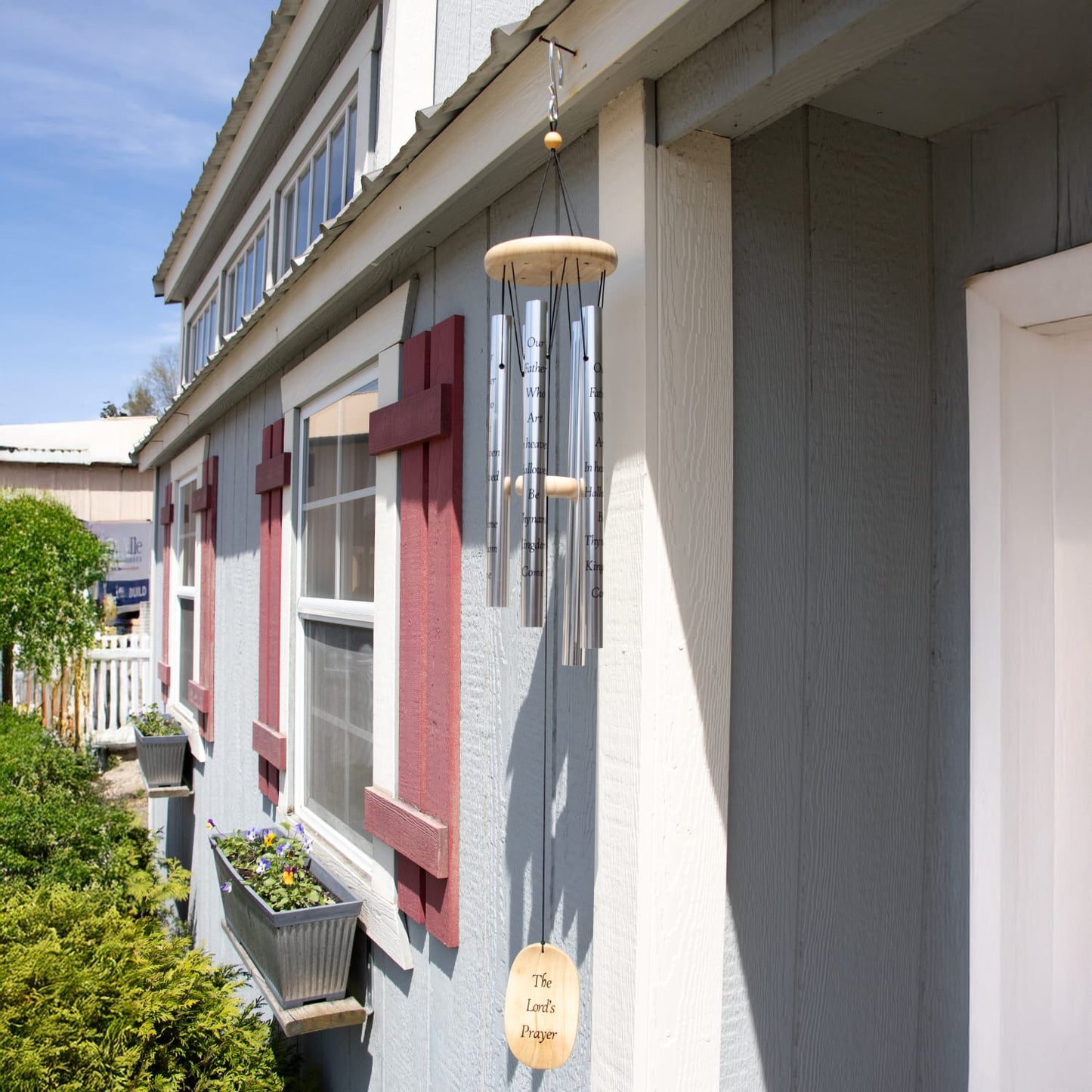 Handcrafted silver windchime with wooden accents hanging from a porch beam against a gray house with red trim, featured in Farm, Garden & Tool and Garden Accessories collections.
