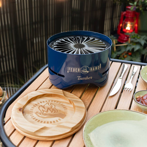 Handcrafted blue charcoal grill with radial vent pattern and logo engraving, set on a wooden table with a carved wooden lid and outdoor dining accessories, perfect for Farm, Garden & Tool, Camping, and Grilling collections.
