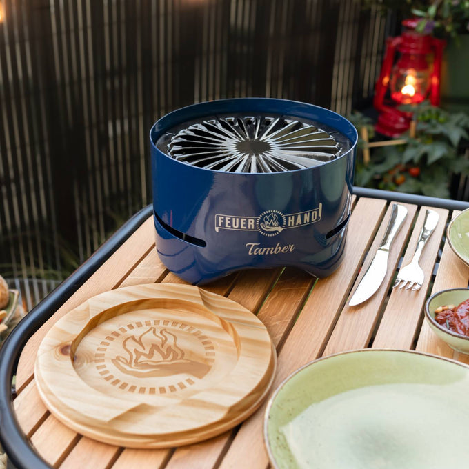 Handcrafted blue charcoal grill with radial vent pattern and logo engraving, set on a wooden table with a carved wooden lid and outdoor dining accessories, perfect for Farm, Garden & Tool, Camping, and Grilling collections.