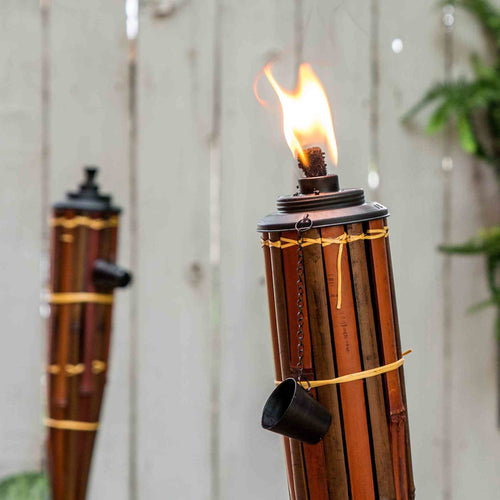 Burning bamboo tiki torch with a metal snuffer cup hanging on the side, set outdoors against a blurred wooden fence and greenery, perfect for Lamps & Lighting and Promo Eligible collections.