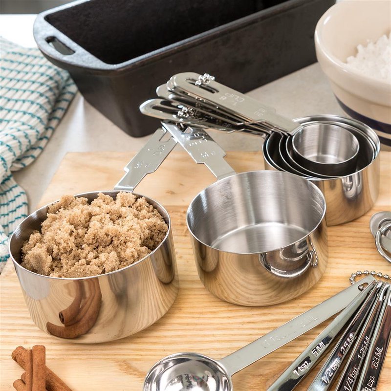 Set of heavy-duty stainless steel measuring cups and spoons with clear well-marked handles, nested neatly on a wooden surface alongside cinnamon sticks and kitchen items in a food prep setting.