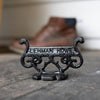 Close-up of a glossy black cast aluminum boot scraper with vintage scroll design and raised lettering, handcrafted by Amish artisans, placed on a rustic floor near a brown boot, ideal for Home & Decor and USA Made collections.