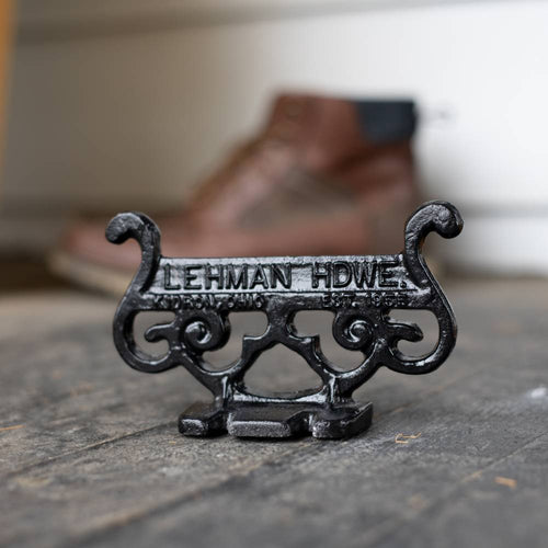 Close-up of a glossy black cast aluminum boot scraper with vintage scroll design and raised lettering, handcrafted by Amish artisans, placed on a rustic floor near a brown boot, ideal for Home & Decor and USA Made collections.