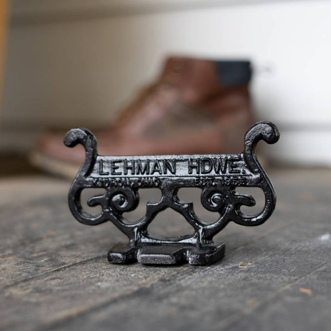 Close-up of a glossy black cast aluminum boot scraper with vintage scroll design and raised lettering, handcrafted by Amish artisans, placed on a rustic floor near a brown boot, ideal for Home & Decor and USA Made collections.