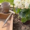 Hand tool with a mirror-polished stainless steel tip and smooth wooden handle resting by a flowerbed and pots, ideal for Farm, Garden & Tool, Garden Accessories, and Seeds and Planting collections.