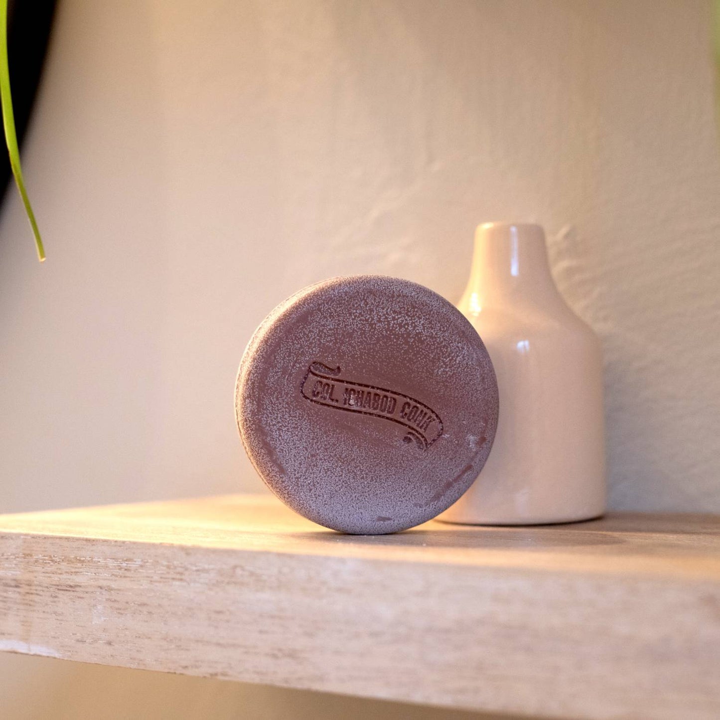 Round handmade shaving soap bar with embossed logo, resting on a wooden shelf next to a simple cream ceramic vase, part of Health & Wellness, Grooming and Soaps collections.