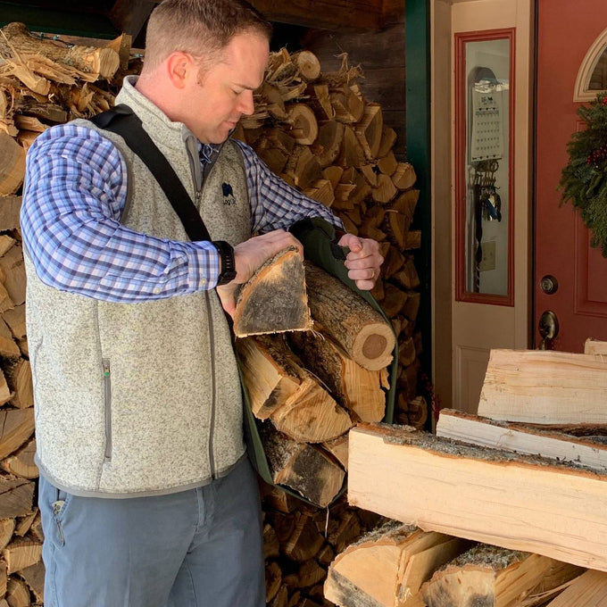 Man using a WoodOX Sling Firewood Carrier with black adjustable straps to lift and hold split firewood near a stacked woodpile, shown outdoors by a red door, suitable for Farm, Garden & Tool, Wood Cutting and Hauling, USA Made, Hearth Accessories, Best Sellers, and Promo Eligible collections.