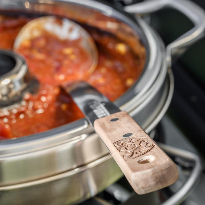 Handcrafted stainless steel serving spoon with a walnut handle featuring an engraved logo, resting on a pot of simmering tomato-based stew, ideal for Farm, Garden & Tool, Kitchen & Food, Camping, Storage and Serving, Best Sellers, Promo Eligible collections.