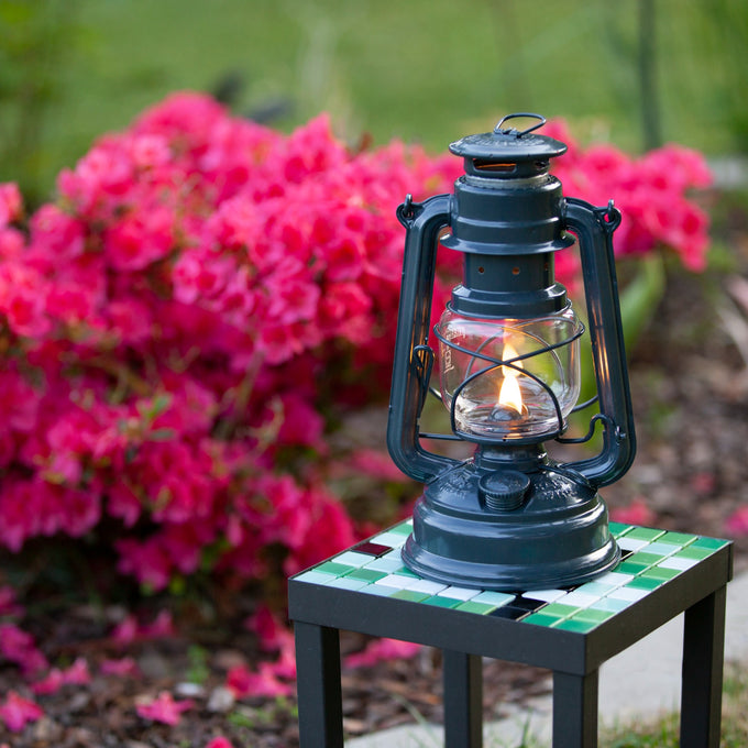 Handcrafted dark galvanized oil lantern with thermal heat-resistant SUPRAX globe, glowing warmly on a tiled outdoor table near vibrant pink flowers, ideal for camping and emergency supplies.