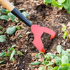 Close-up of a hand tool with a red steel head and wooden handle digging into soil among green plants, handcrafted and Amish made, featured in Farm, Garden & Tool and USA Made collections.