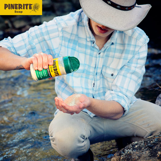 Person wearing a light plaid shirt and wide-brimmed hat pouring granular all-natural pine soap onto their hand near a river, featured in Health & Wellness, Soaps, and USA Made collections.