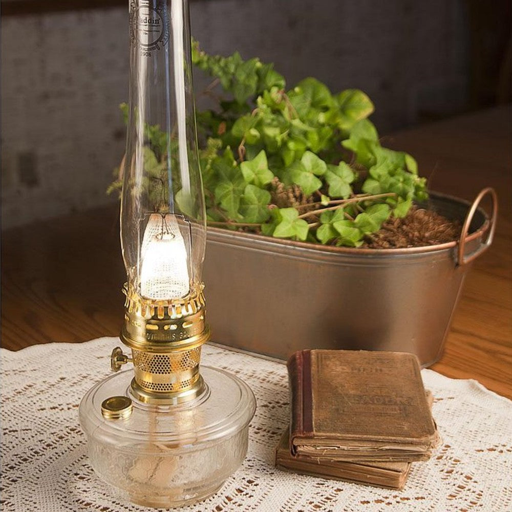 Glass oil lamp with brass fittings glowing softly on a lace doily beside worn vintage books and a metal planter of green ivy, perfect for Lamps & Lighting and Lamp and Lantern Fuel collections.