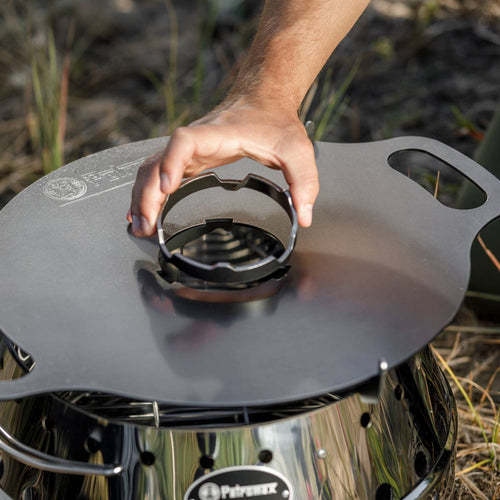 Hand placing a round metal cooking support piece into the center opening of a matte black griddle plate atop a stainless steel camping stove, suitable for outdoor grilling in Farm, Garden & Tool, Stoves, Camping, and Grilling collections.