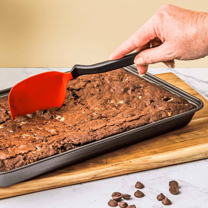 Hand using a flexible red spatula with a black handle to cut or lift freshly baked chocolate brownies in a rectangular baking pan, set on a wooden board with chocolate chips nearby, ideal for Kitchen & Food, Kitchen Gadgets, Cooking and Baking Helpers, Best Sellers, and Promo Eligible collections.