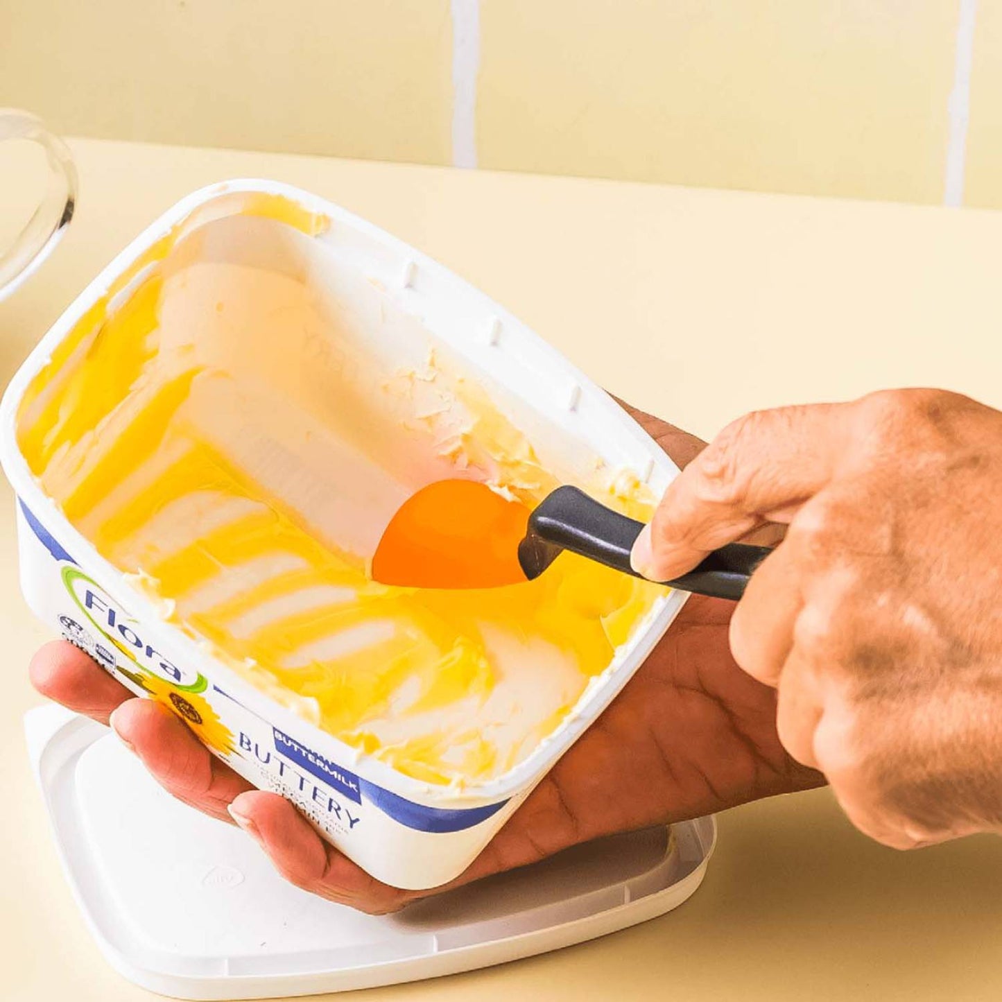 Person using a flexible orange silicone spoon with black handle to scrape buttery spread from a rectangular plastic tub, showcasing a useful tool for Kitchen & Food, Food Prep, and Measuring collections.