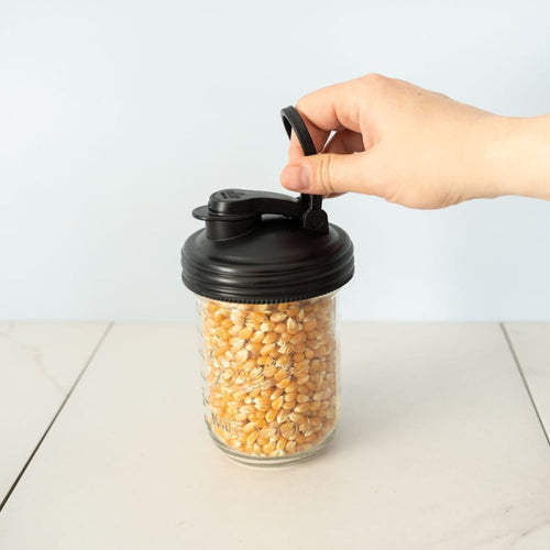 Hand lifting a black pour lid with a carry loop and flip-top spout on a glass jar filled with yellow corn kernels, ideal for Kitchen & Food, Kitchen Gadgets, USA Made, and Best Sellers collections.