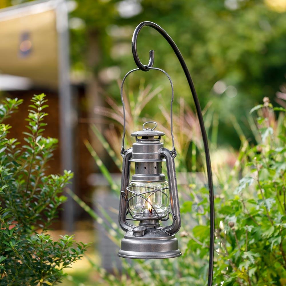 Black metal lantern holder with curved hook displaying a lit lantern outdoors among green foliage, suitable for Lamps & Lighting, Camping, and Emergency Supplies collections.