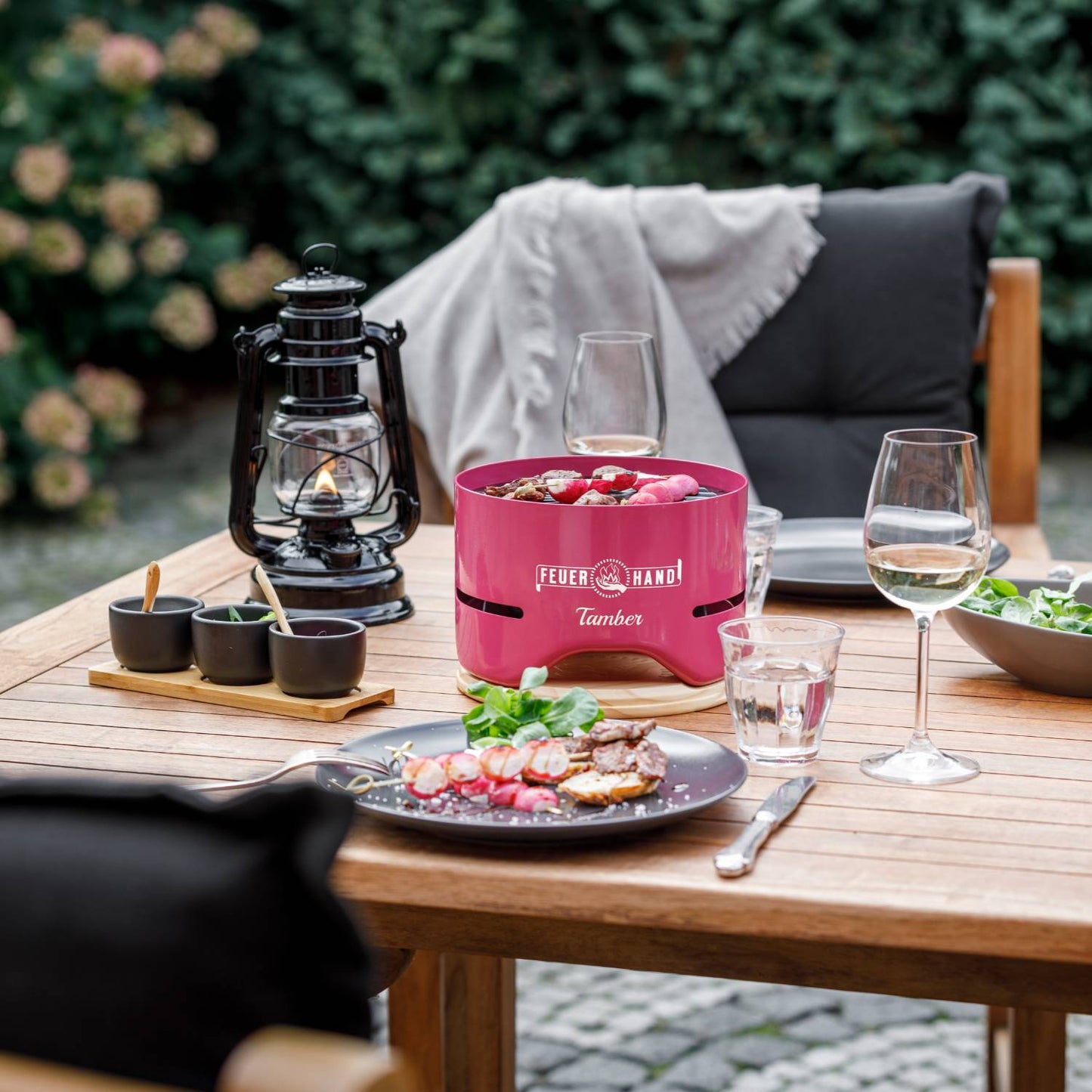Handcrafted pink tabletop charcoal grill cooking small skewers, set on a wooden outdoor dining table with glassware and a black vintage lantern, ideal for Farm, Garden & Tool and Grilling collections.