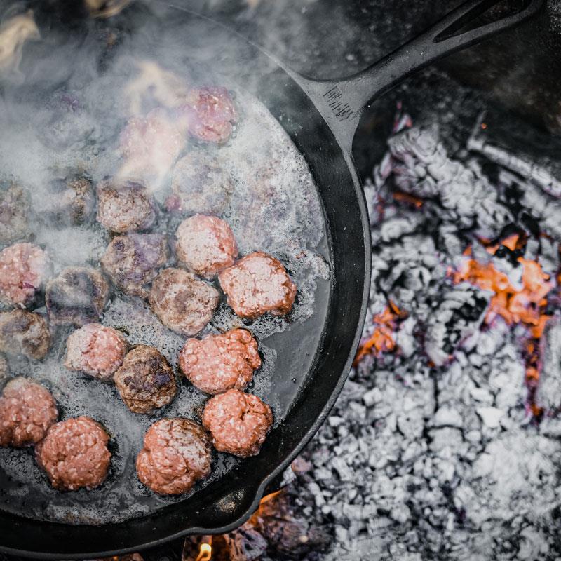 Cast iron fire skillet with long handle cooking meat patties over glowing embers, showcasing durable craftsmanship for Farm, Garden & Tool, Kitchen & Food, Camping, Cookware, Cast Iron, Best Sellers, and Promo Eligible collections.