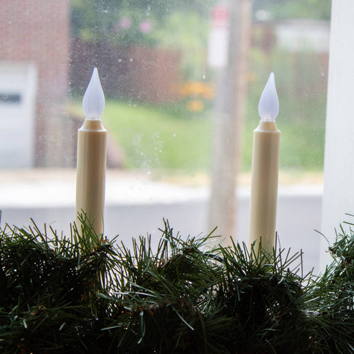 Pair of cordless LED taper candles with amber flickering flames atop slim bases nestled in green garland on a windowsill, perfect for holiday decor and best sellers collections.