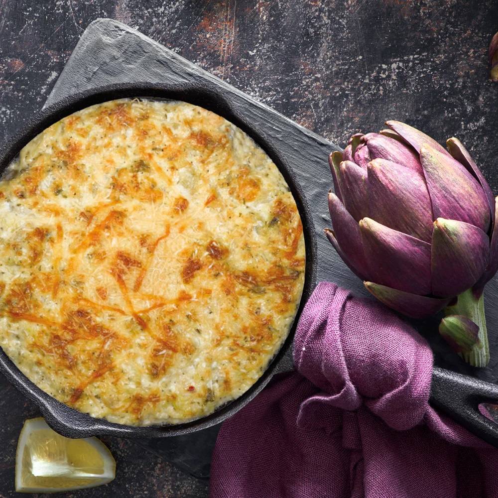 Baked creamy artichoke dip with golden melted cheese in an 8-inch cast iron skillet, styled with fresh artichoke, lemon wedge, and purple cloth, perfect for Kitchen & Food, Dry Goods and Mixes, Best Sellers, Promo Eligible collections.